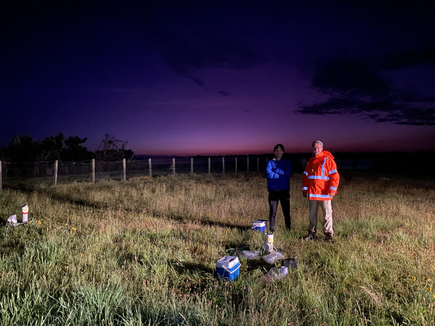 Two people stand under a dusky sky in a field, near scientific sampling gear